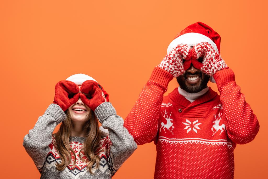 feliz casal interracial em santa chapéus, mitenes e camisolas de Natal imitando binóculos com as mãos no fundo laranja
 - Foto, Imagem