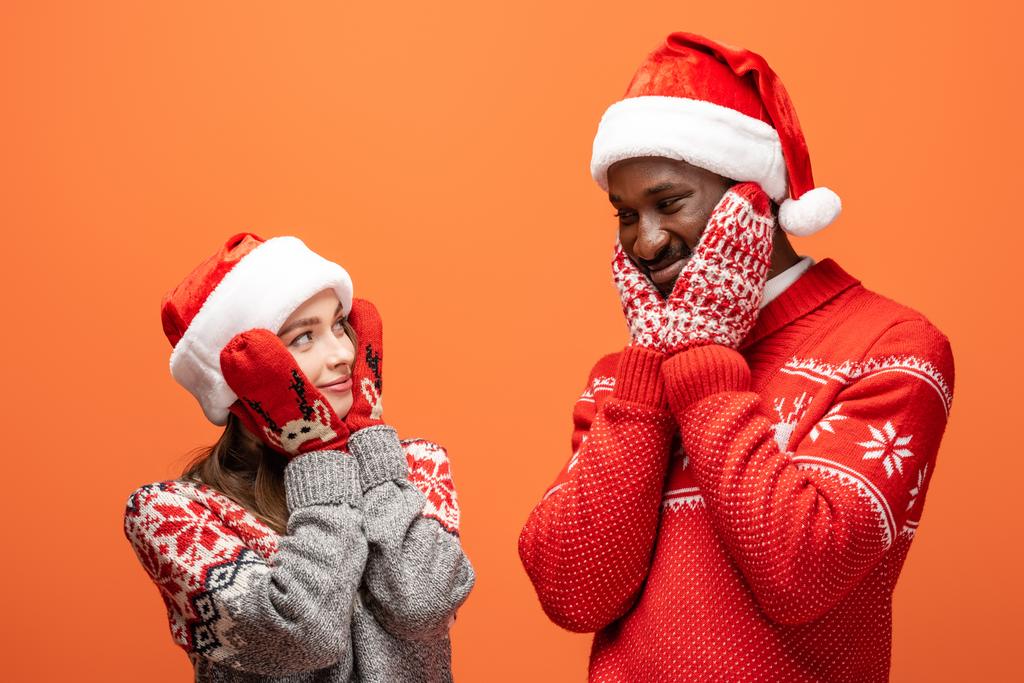 onnellinen rotujenvälinen pari santa hatut, lapaset ja joulun villapaidat koskettaa posket oranssi tausta
 - Valokuva, kuva