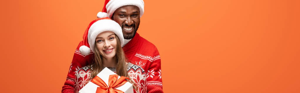 happy interracial couple in santa hats and Christmas sweaters hugging and holding gift on orange background, panoramic shot - Photo, Image