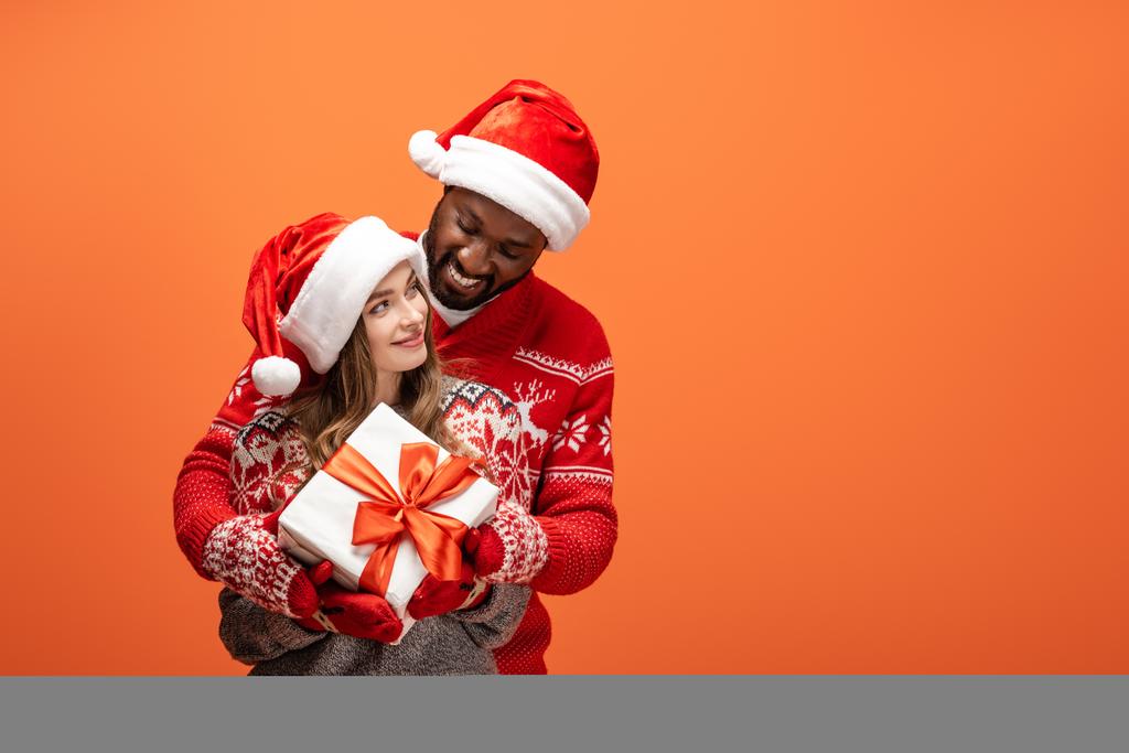 ευτυχισμένο διαφυλετικό ζευγάρι σε καπέλα Σάντα και πουλόβερ Χριστουγέννων αγκαλιάζει και κρατώντας δώρο σε πορτοκαλί φόντο - Φωτογραφία, εικόνα