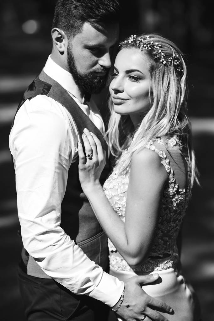 Sensual Black and white foto of bride and groom. Stylish couple of happy newlyweds posing in the park on their wedding day. Handsome bearded groom admires and kisses pretty bride. Together. - Photo, Image