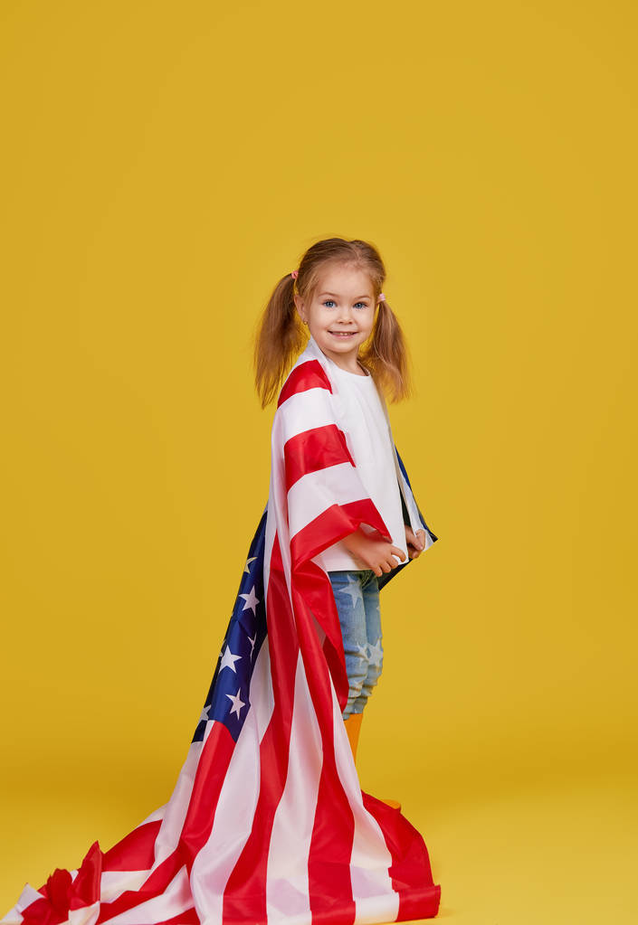 Vacaciones patrióticas. Niño feliz, linda niña con bandera americana en el fondo del estudio amarillo. EE.UU. celebra el 4 de julio, Día de la Independencia
. - Foto, imagen