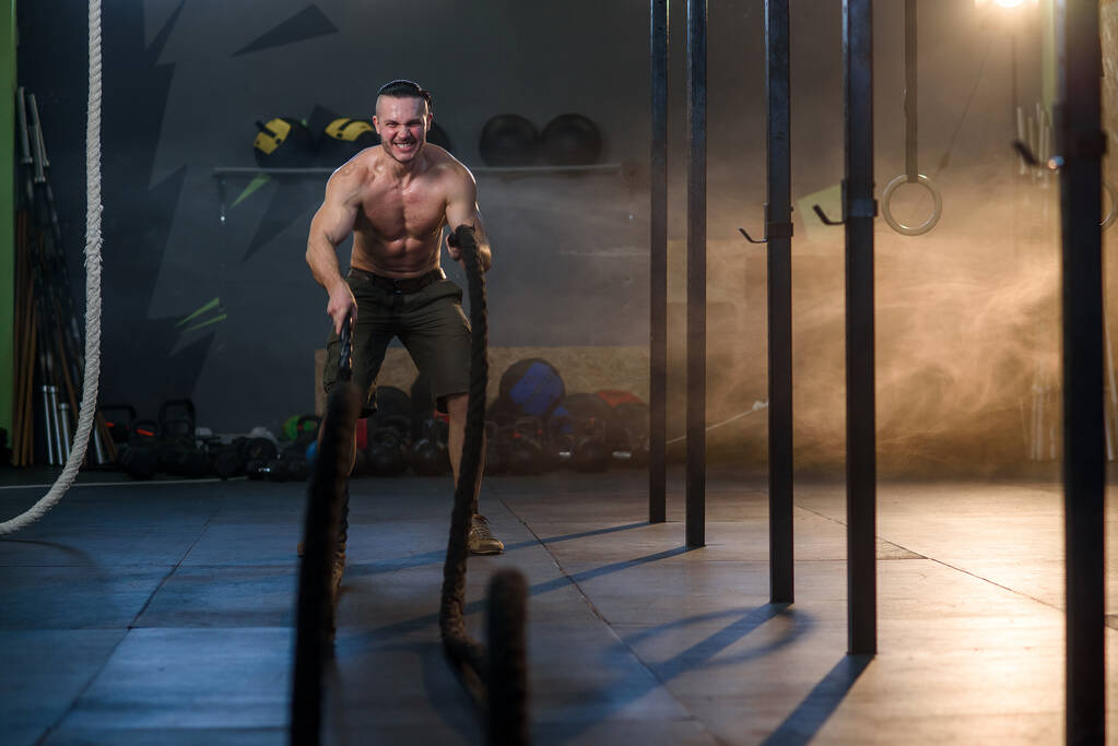 Όμορφος ισχυρός άνθρωπος κάνει ασκήσεις για τους μυς με σχοινιά μάχης στο γυμναστήριο. - Φωτογραφία, εικόνα