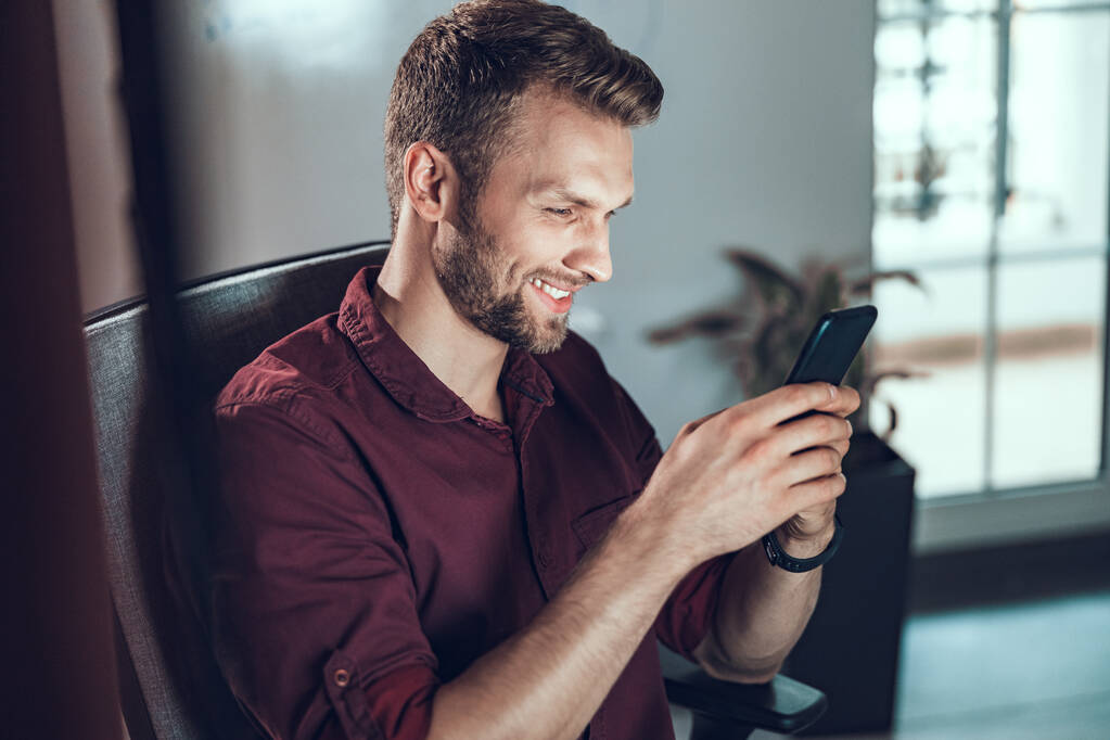 Usmívající se mladý muž dívá na obrazovce telefonu v kanceláři - Fotografie, Obrázek