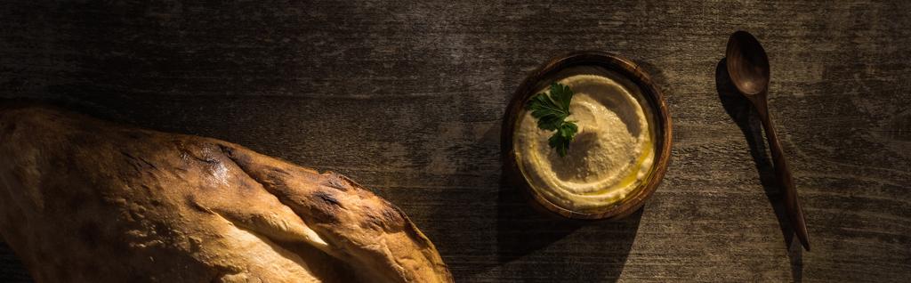 Draufsicht auf köstlichen Hummus in Schüssel in der Nähe von frisch gebackener Flaka und Löffel auf rustikalem Holztisch, Panoramaaufnahme - Foto, Bild