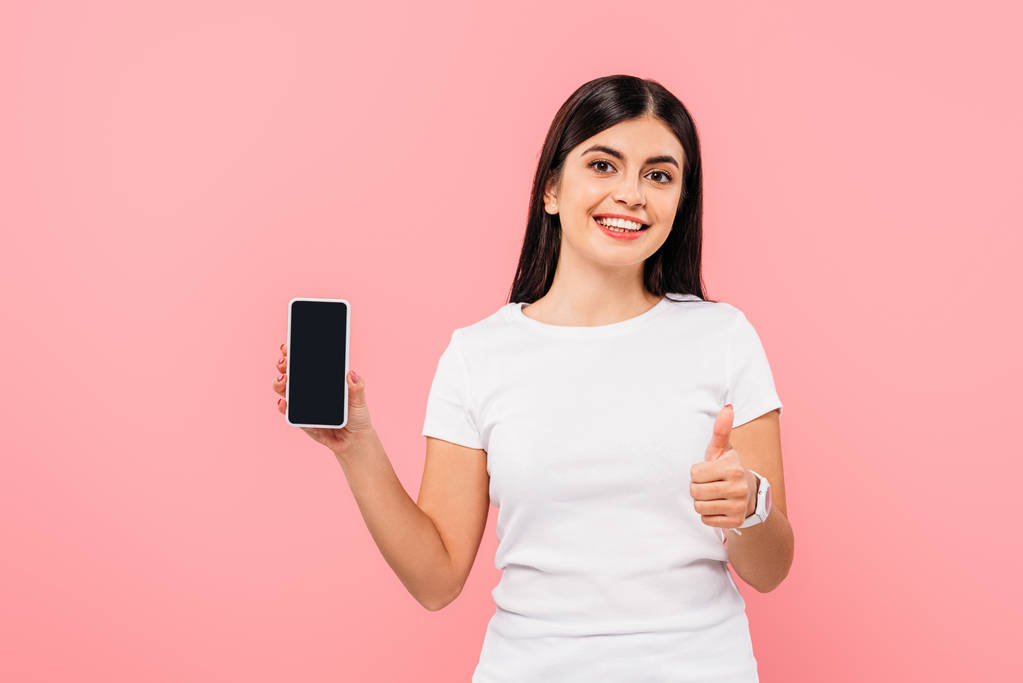 usmívající se hezká brunetka dívka drží smartphone s prázdnou obrazovkou a ukazující palec nahoru izolované na růžové - Fotografie, Obrázek