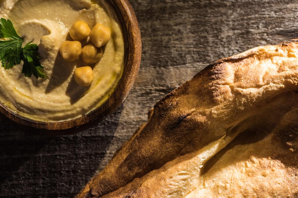 vista superior del delicioso hummus con garbanzos en un tazón cerca de la pita recién horneada sobre una mesa rústica de madera
 - Foto, Imagen