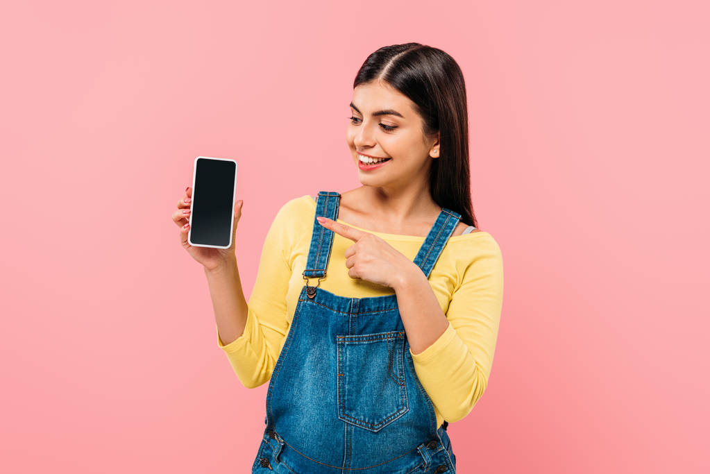 улыбающаяся беременная девушка, указывающая пальцем на смартфон с пустым экраном, изолированным на розовом
 - Фото, изображение