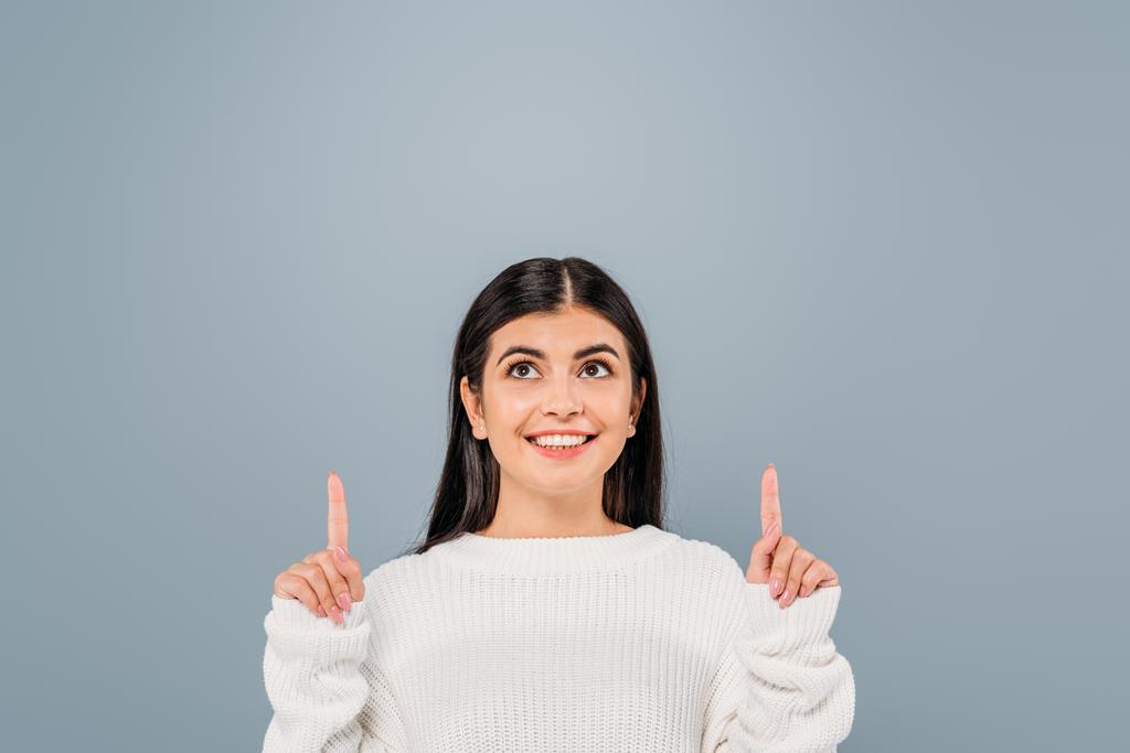 smiling pretty brunette girl in white sweater pointing with fingers upwards isolated on grey - Photo, Image