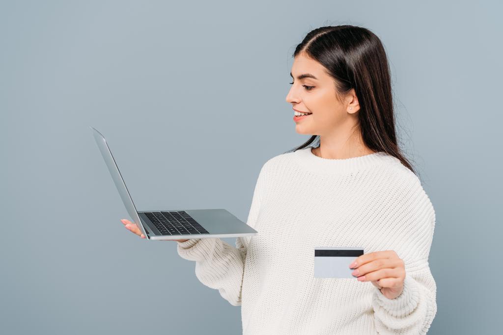 sonriente chica bonita en suéter blanco sosteniendo portátil y tarjeta de crédito aislado en gris
 - Foto, Imagen