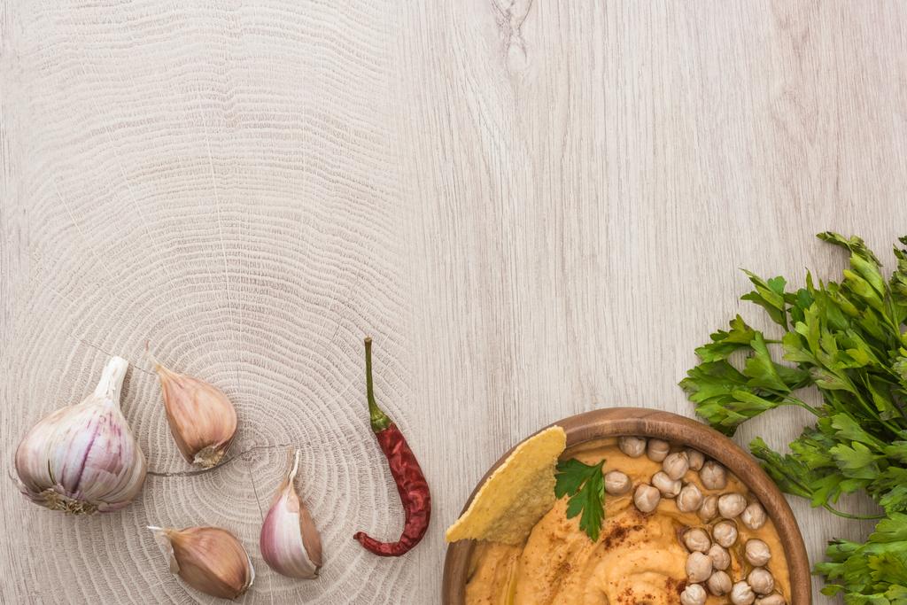 Draufsicht auf köstlichen Hummus mit Kichererbsen, Nacho in Schüssel in der Nähe von Gewürzen und Petersilie auf beigem Holztisch - Foto, Bild