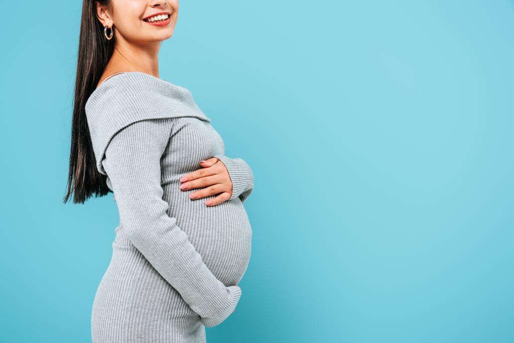 vista lateral de sorrir grávida bonita menina tocando barriga isolada no azul
 - Foto, Imagem