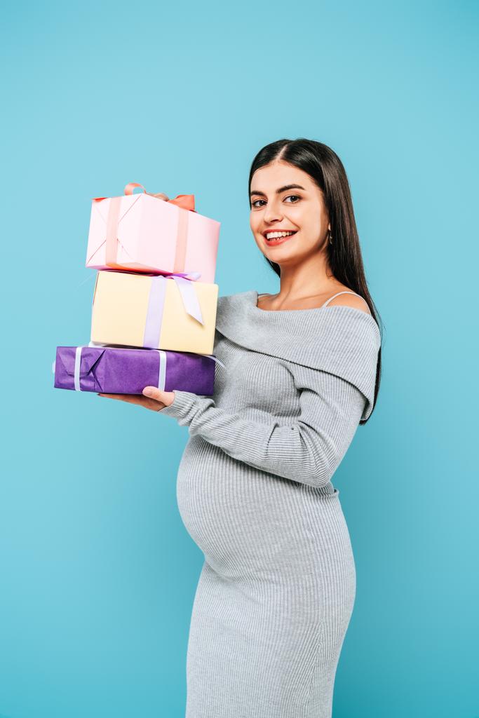 Улыбающаяся беременная красивая девушка держит подарки изолированы на синий
 - Фото, изображение