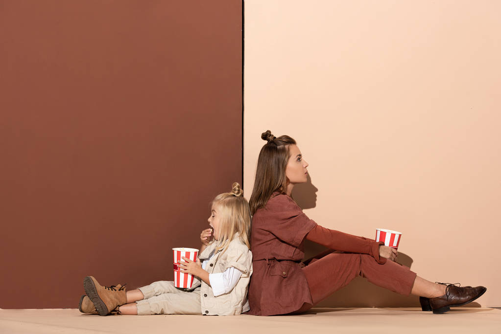 zurück nach hinten Ansicht der Tochter beim Popcornessen und der attraktiven Mutter auf beigem und braunem Hintergrund  - Foto, Bild
