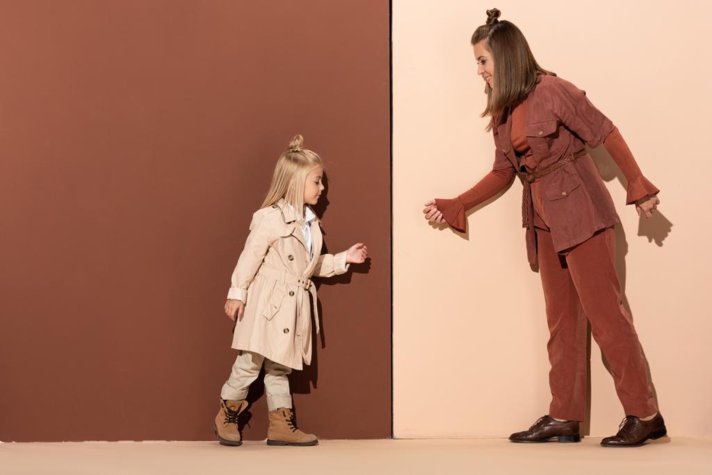 zijaanzicht van dochter en lachende moeder lopen op beige en bruine achtergrond  - Foto, afbeelding