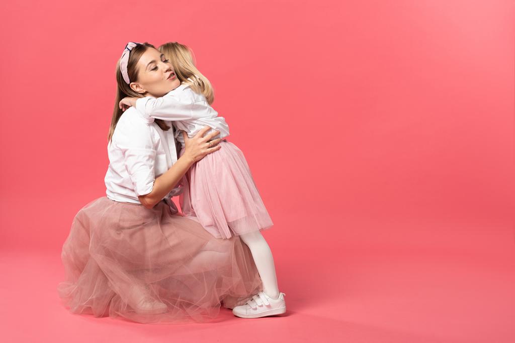 hija abrazando atractiva y sonriente madre sobre fondo rosa
  - Foto, imagen