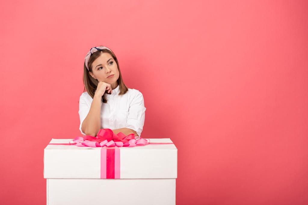 mulher atraente e pensativo de pé perto caixa de presente isolado em rosa
 - Foto, Imagem