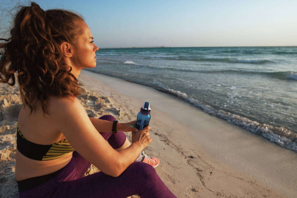 jeune femme sportive en vêtements de sport avec bouteille d'eau regardant au loin tout en étant assis sur le bord de la mer au coucher du soleil
. - Photo, image