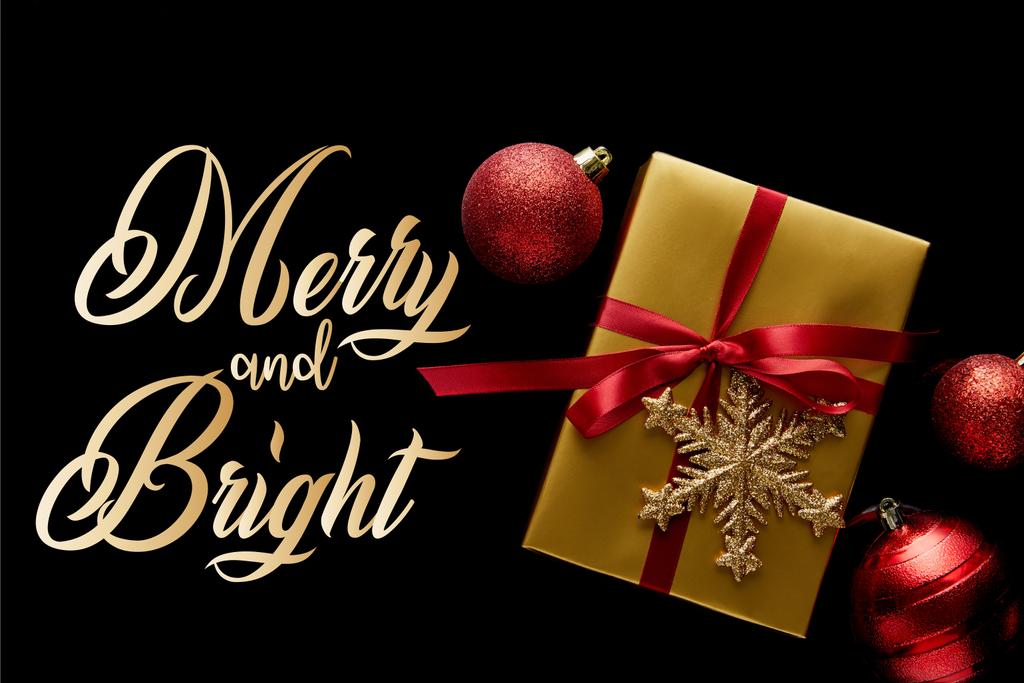 Kırmızı kurdeleli ve mücevherlerin yakınındaki kar taneli parlak altın Noel hediyesinin en üst görüntüsü. Siyah üzerine izole edilmiş, neşeli ve parlak bir resimle. - Fotoğraf, Görsel