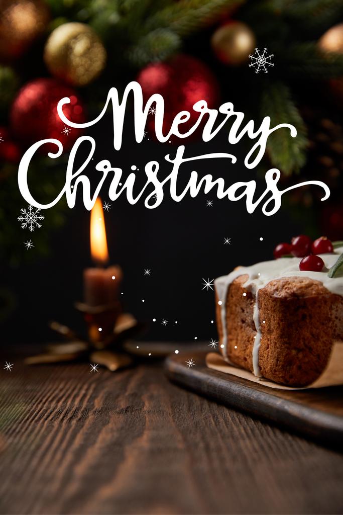 selective focus of traditional Christmas cake with cranberry near Christmas wreath with baubles and burning candle on wooden table with Merry Christmas illustration - Photo, Image