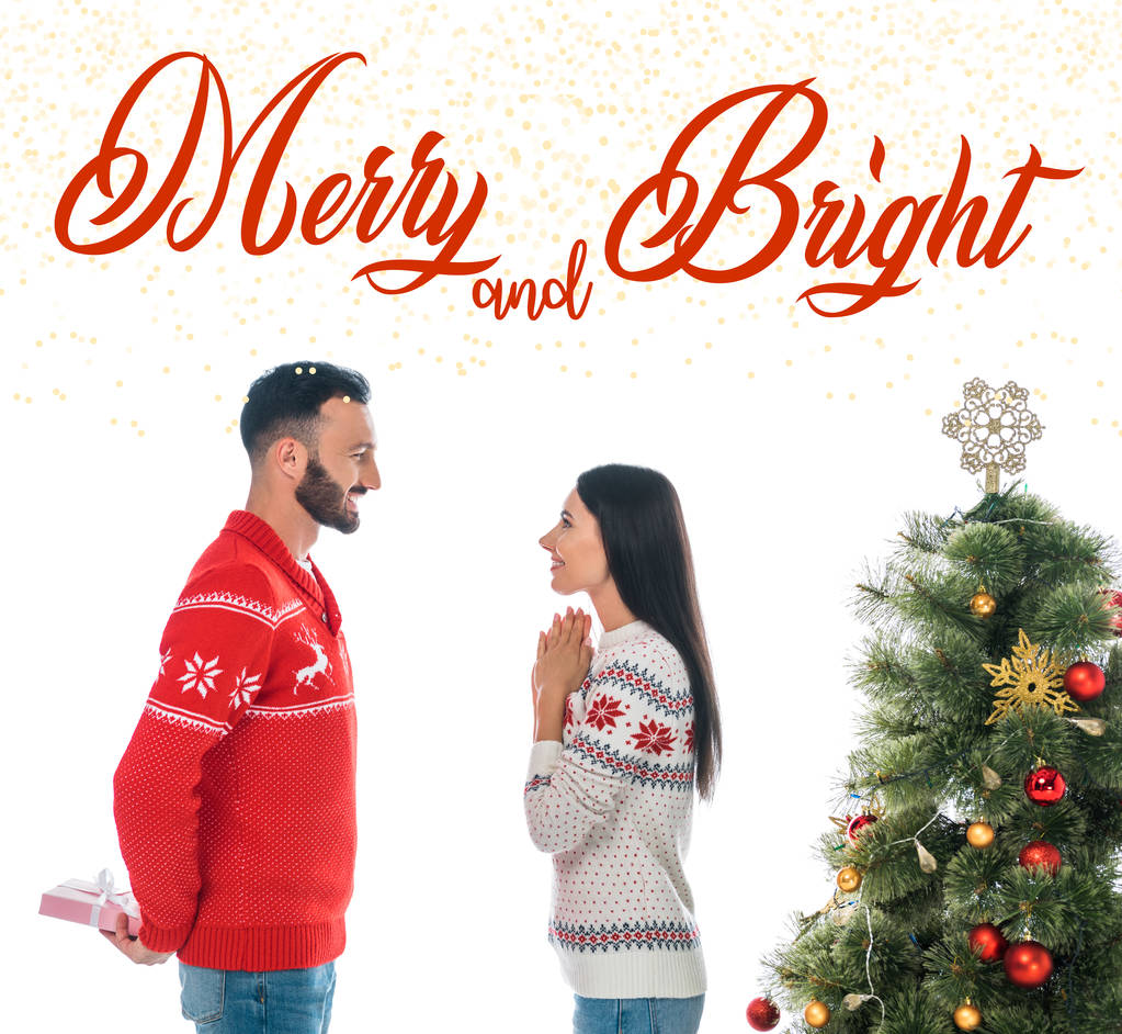 happy bearded man holding gift near woman and christmas tree isolated on white with merry and bright lettering - 写真・画像