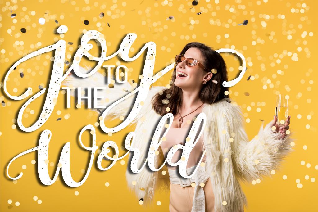 Sahte kürk ceketli ve güneş gözlüklü heyecanlı parti kızı konfeti altında, sarıda izole edilmiş şampanyayla dünya resmine neşe katıyor. - Fotoğraf, Görsel