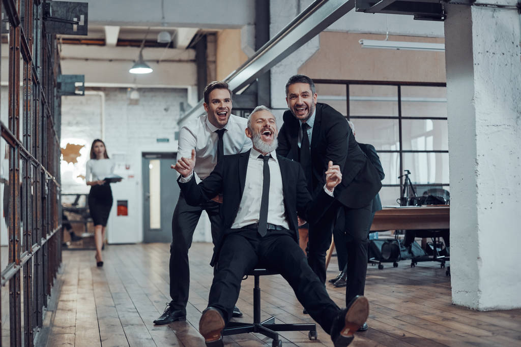 Empresários alegres empurrando seu chefe na cadeira de escritório enquanto correm no corredor
 - Foto, Imagem