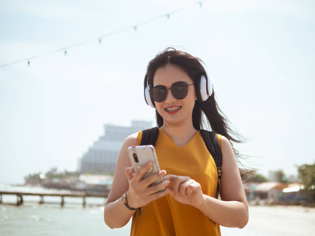 Mujer asiática feliz con mochila usando auriculares en la playa, li
 - Foto, Imagen