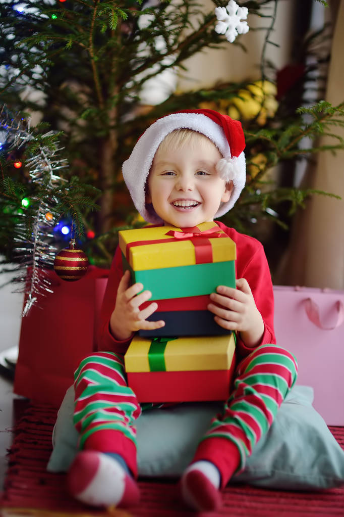 自宅でモミの木の下でクリスマスプレゼントを持つ興奮した小さな男の子。クリスマスの朝の幸せな子供の肖像画. - 写真・画像