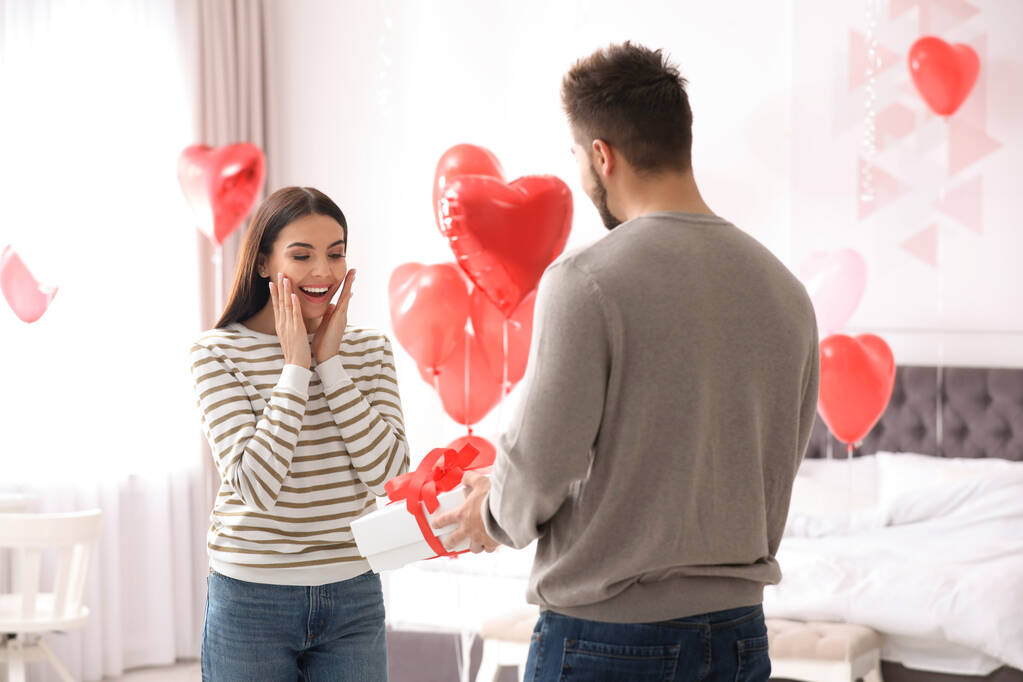 Hombre joven presentando regalo a su novia en el dormitorio decorado con globos en forma de corazón. Celebración de San Valentín
 - Foto, imagen