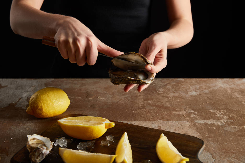 обрезанный вид женщины с ножом во время открытия устрицы рядом с лимонами изолированы на черном
  - Фото, изображение