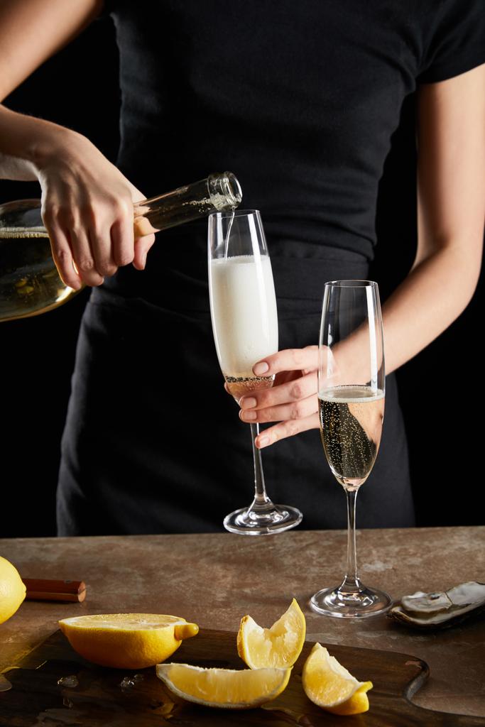 Selektiver Fokus der Frau, die Sekt in Champagnerglas in der Nähe von Austern und Zitronen einschenkt, isoliert auf Schwarz  - Foto, Bild