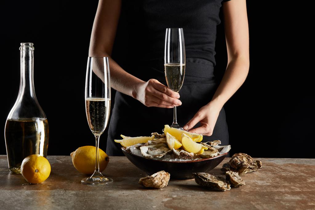 黒で隔離された氷のボウルにカキやレモンの近くに輝くワインとガラスを保持する女性のクロップドビュー  - 写真・画像