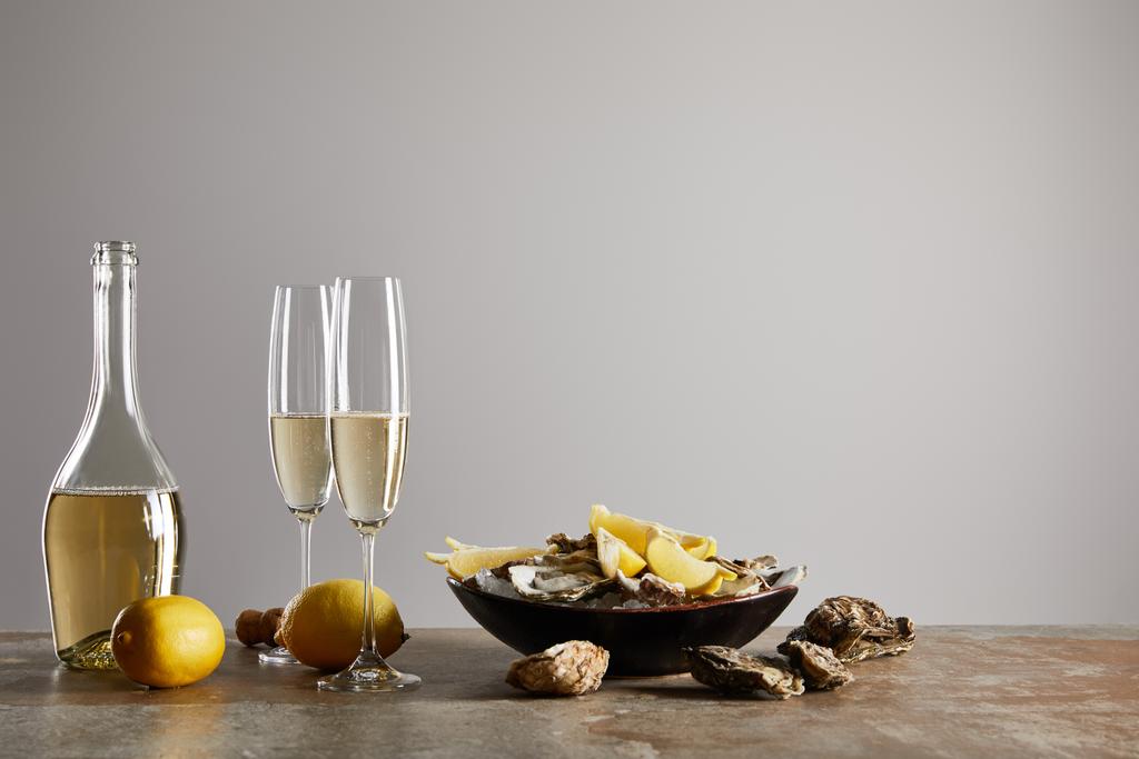 sklenice na šampaňské se šumivým vínem v blízkosti ústřic a citrónů v misce izolované na šedi  - Fotografie, Obrázek