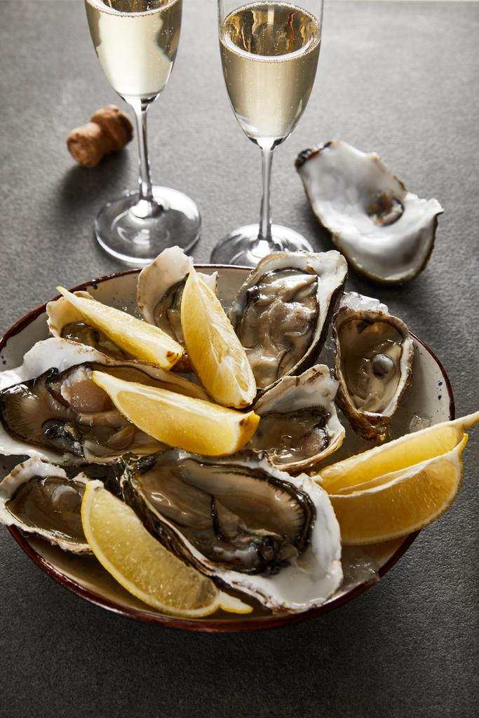 selectieve focus van heerlijke oesters en citroenen in kom bij champagneglazen met mousserende wijn op grijze ondergrond - Foto, afbeelding
