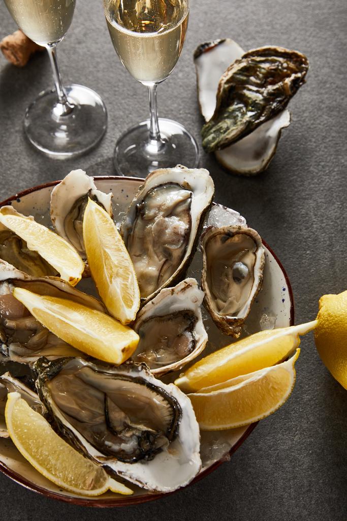 bovenaanzicht van heerlijke oesters en citroenen in kom bij champagneglazen met mousserende wijn op grijze ondergrond - Foto, afbeelding