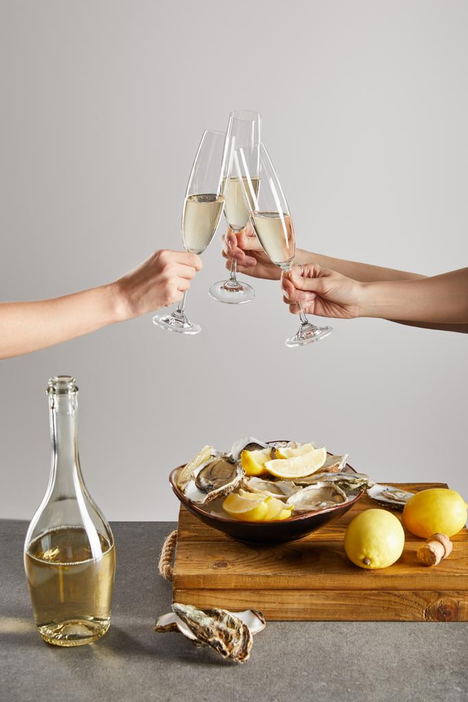 bijgesneden beeld van vrouwen die champagneglazen met mousserende wijn in de buurt van kom met oesters en citroenen geïsoleerd op grijs  - Foto, afbeelding