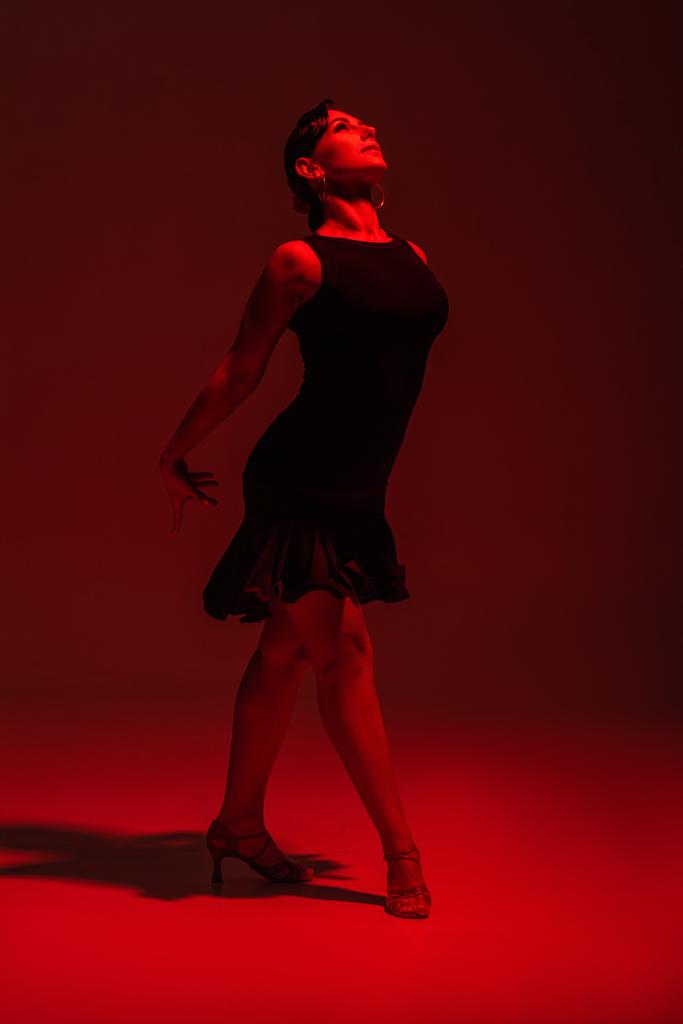 beautiful dancer in black dress performing tango on dark background with red lighting - Photo, Image
