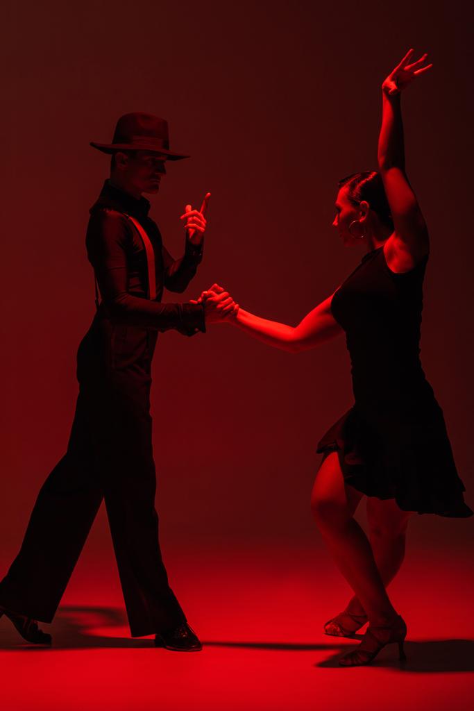 elegant couple of dancers in black clothing performing tango on dark background with red illumination - Photo, Image