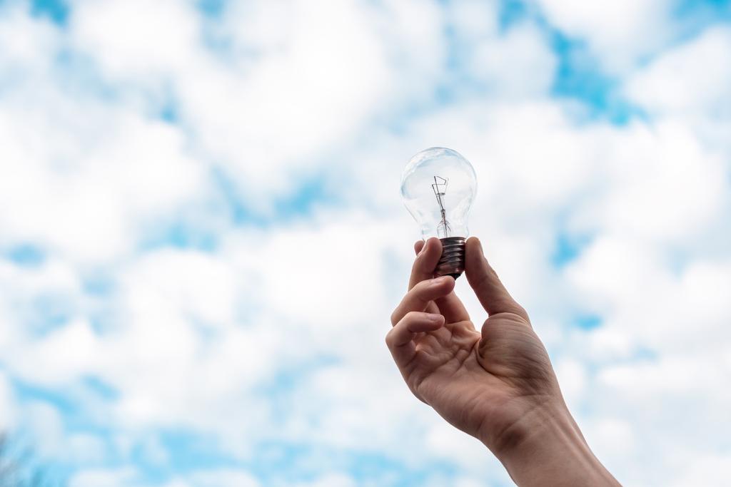 cropped view of woman holding light bulb with blue sky and clouds on background - Photo, Image