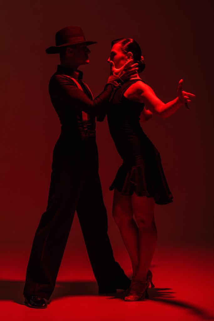 pareja expresiva de bailarines en ropa negra realizando tango sobre fondo oscuro con iluminación roja
 - Foto, Imagen