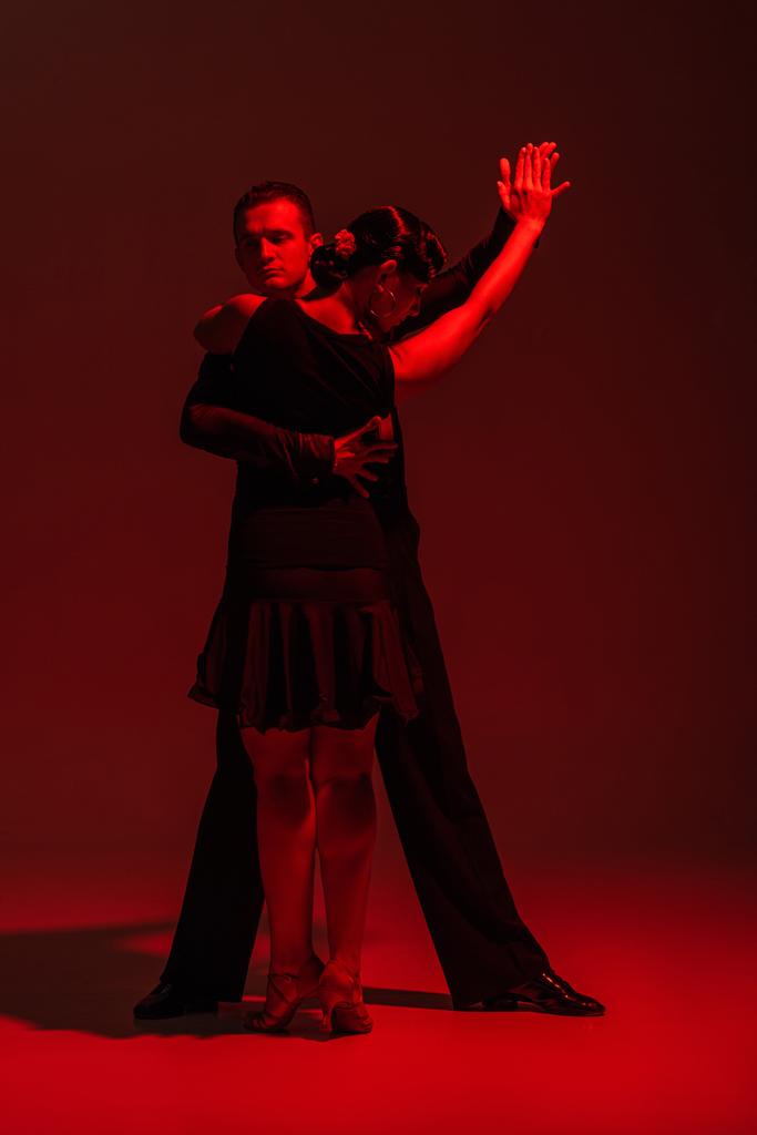 elegant couple of dancers in black clothing performing tango on dark background with red lighting - Photo, Image
