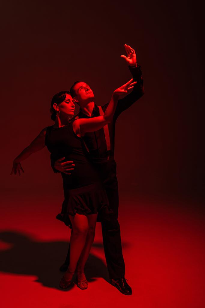 стоїть англійська пара танцюристів в чорному одязі, виконуючи танго на темному фоні з червоним освітленням
 - Фото, зображення