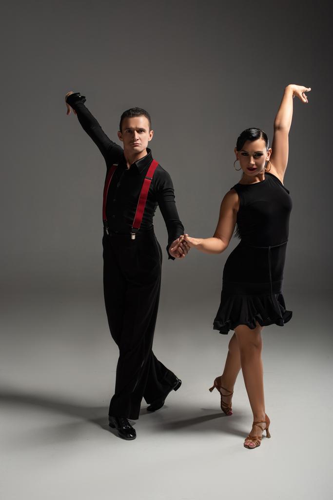 elegantní tanečníci v černém oblečení při pohledu na kameru při tanci tanga na šedém pozadí - Fotografie, Obrázek