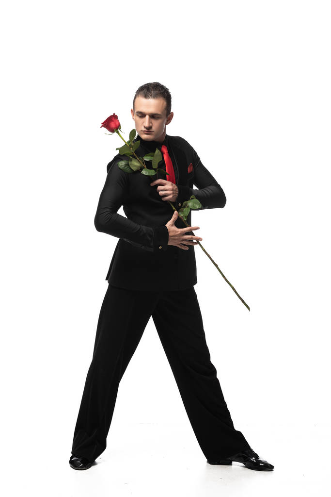 sebevědomý, smyslný tanečník tanga v elegantním černém obleku držící červenou růži na bílém pozadí - Fotografie, Obrázek