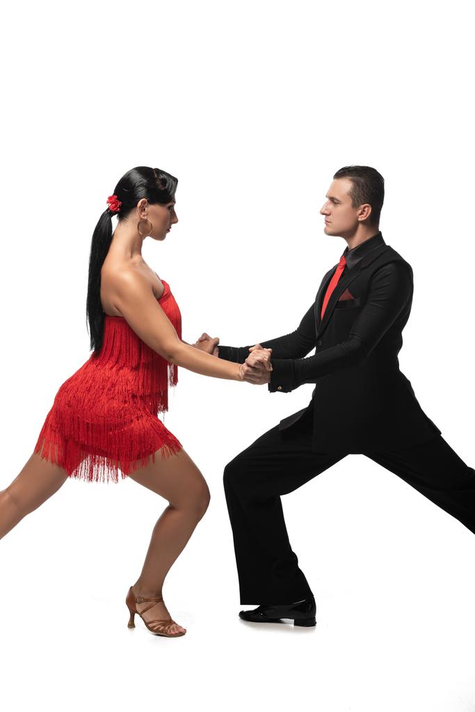passionele, elegante dansers die elkaar aankijken en elkaars hand vasthouden terwijl ze tango op een witte achtergrond uitvoeren - Foto, afbeelding