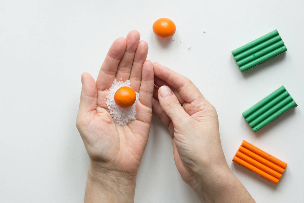 Kleine oranje bal gemaakt van polymeer klei op vrouw hand met zeezout te maken deuken op oranje fruit op witte achtergrond - Foto, afbeelding