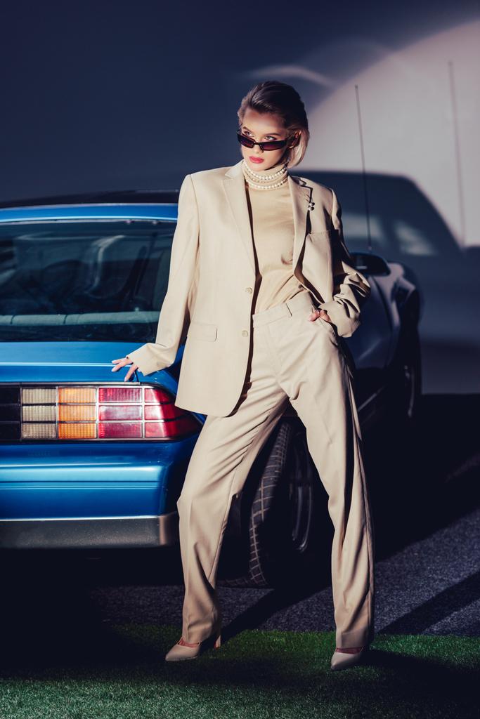 Eski arabanın yanında duran takım elbiseli ve güneş gözlüklü çekici ve şık bir kadın.  - Fotoğraf, Görsel