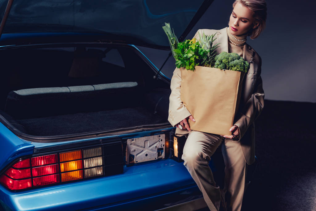aantrekkelijke en stijlvolle vrouw in pak staan in de buurt van retro auto en houden papieren zak met voedsel  - Foto, afbeelding