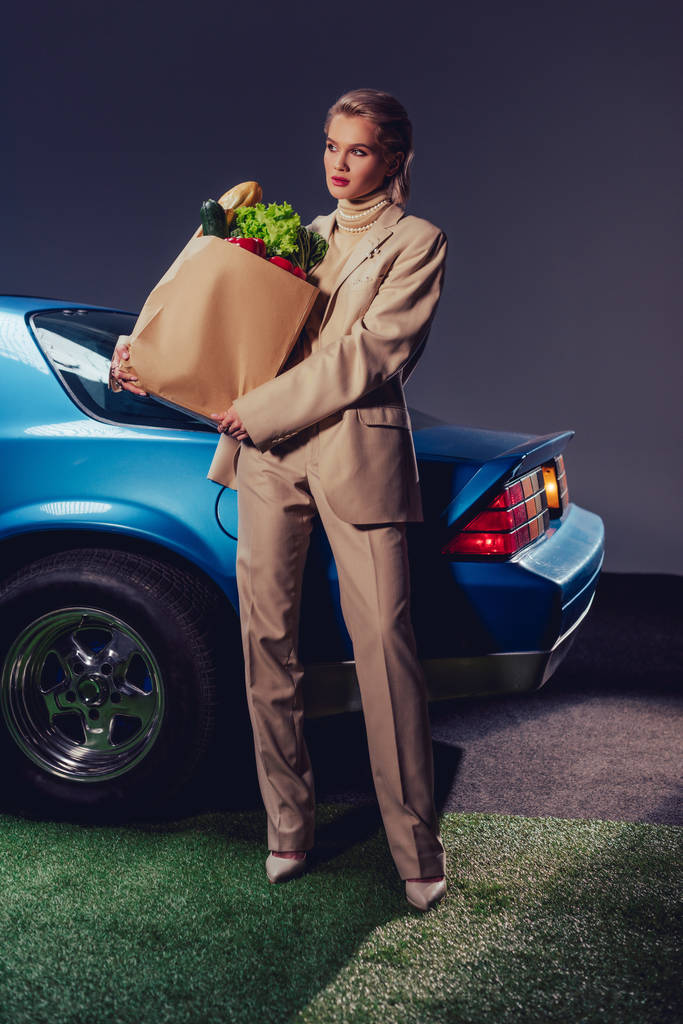 attraktive und stilvolle Frau im Anzug, die neben dem Retro-Auto steht und Papiertüten mit Lebensmitteln in der Hand hält  - Foto, Bild